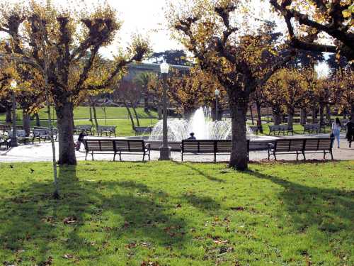 A park scene with benches, autumn trees, and a fountain surrounded by green grass and sunlight.