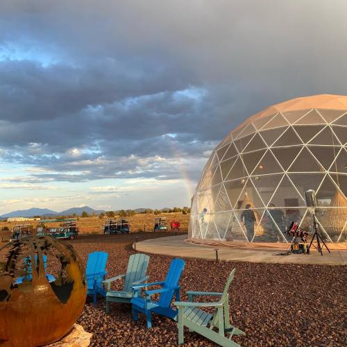 A geodesic dome structure with colorful chairs outside, set against a cloudy sky and distant mountains, with a rainbow visible.