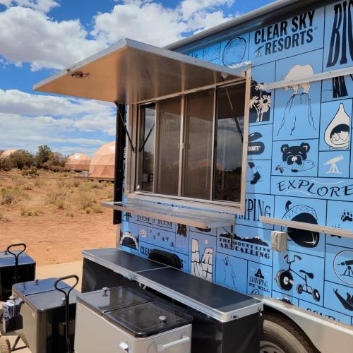 A food truck with a blue exterior featuring various outdoor-themed graphics, set in a desert landscape with domes in the background.