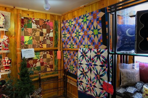 Colorful quilts displayed on wooden walls in a cozy shop, with a small tree and decorative items nearby.