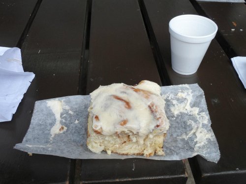 A cinnamon roll with cream cheese frosting on a paper wrapper, next to a white cup, on a wooden table.