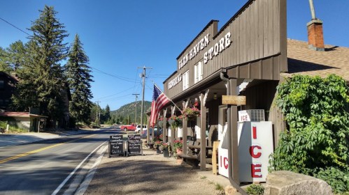 A rustic general store with an American flag, surrounded by trees and mountains, along a quiet road.