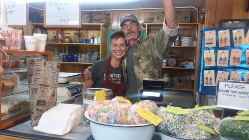 A smiling couple stands behind a counter filled with colorful treats, with shelves of products in the background.