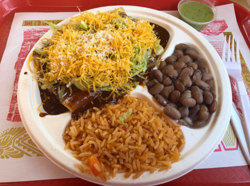 A plate of enchiladas topped with cheese and lettuce, served with rice, pinto beans, and a side of green sauce.