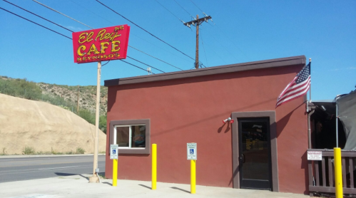 Exterior of El Rey Café with a red building, American flag, and yellow poles, set against a clear blue sky.