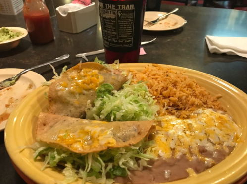 A plate of tacos topped with cheese and lettuce, served with rice and refried beans, with hot sauce in the background.