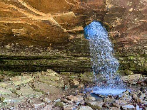 A waterfall cascades from a rock overhang into a rocky area below, surrounded by natural stone formations.