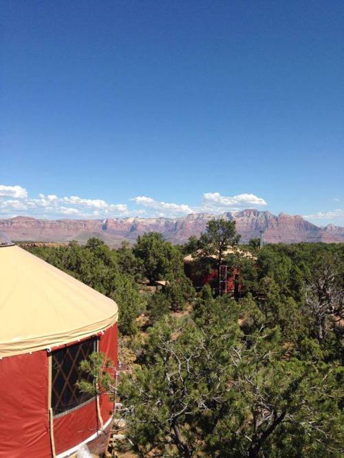 A scenic view of red rock formations and blue sky, with a yurt and trees in the foreground.