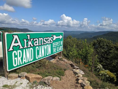 Sign for Arkansas' Grand Canyon, indicating it's the deepest in the Ozarks, with a scenic view of mountains and clouds.