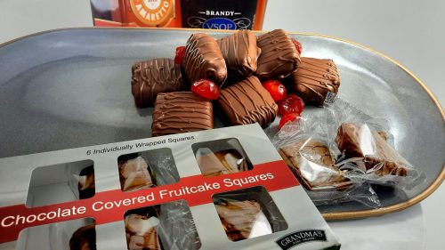 A plate of chocolate-covered fruitcake squares with cherries, next to a box of individually wrapped treats.