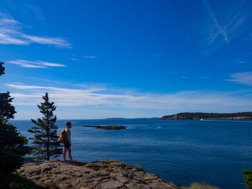 A person stands on a rocky shore, gazing at a calm blue sea under a clear sky with scattered clouds.