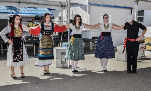 Five dancers in traditional attire perform a folk dance under a tent, holding hands and smiling.