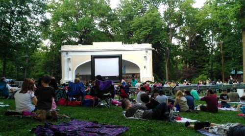 A crowd sits on the grass with blankets and chairs, watching a movie on a large screen in a park.
