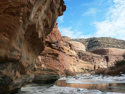 A scenic view of red rock formations beside a frozen river, with blue skies and distant hills in the background.