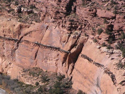 A wooden bridge spans a rocky cliff, with shadows cast on the red rock surface below. Lush greenery is visible nearby.