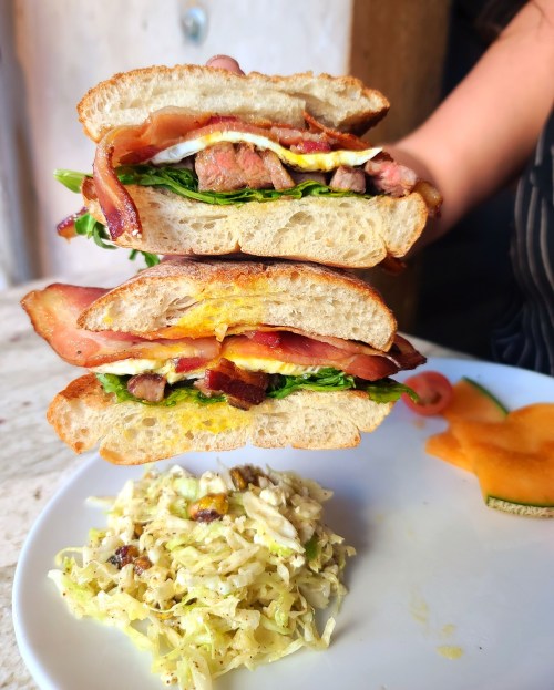 A stacked sandwich with bacon, lettuce, egg, and meat, served with a side of coleslaw and melon on a plate.