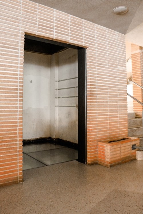 An empty elevator shaft with brick walls and a staircase nearby, featuring a clean floor and minimal lighting.