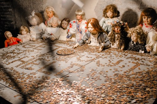 A dimly lit room with dolls surrounding a table covered in coins, spelling out "HELP" in the center.