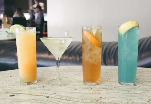 Four colorful cocktails on a marble table: orange, martini, iced tea, and blue drink, with garnishes of lime and orange.