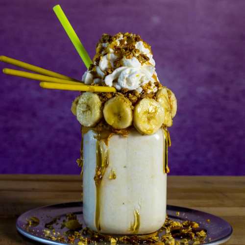 A tall milkshake topped with whipped cream, banana slices, granola, and drizzled with syrup, served with colorful straws.