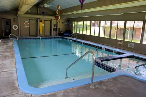 Indoor swimming pool with a hot tub, surrounded by large windows and a clean, tiled deck.