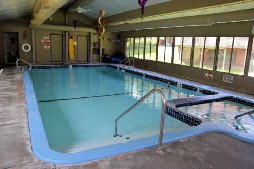 Indoor swimming pool with a hot tub, surrounded by large windows and a clean, tiled deck.