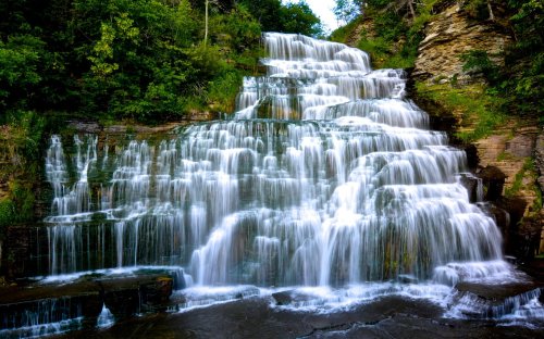 A cascading waterfall flows over rocky steps, surrounded by lush greenery and trees.