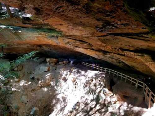 A rocky cave with a wooden railing, sunlight illuminating the ground and people exploring inside.