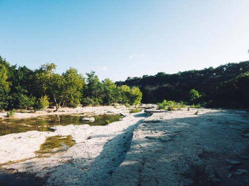 A serene landscape featuring a rocky riverbank, lush greenery, and a clear blue sky.