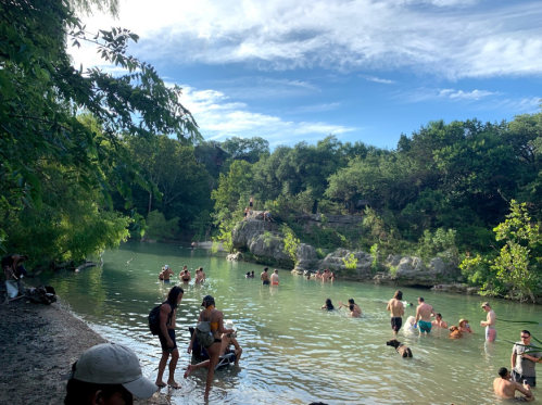 A scenic river with people swimming and relaxing, surrounded by lush greenery and rocky formations under a blue sky.