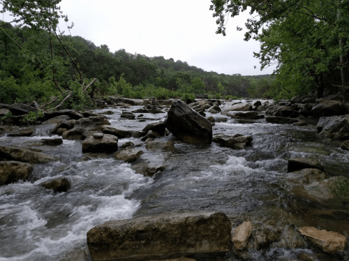 A flowing river with rocky banks, surrounded by lush green trees under a cloudy sky.