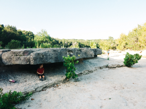 A child sits under a large rock formation in a dry, sandy area surrounded by greenery and trees.