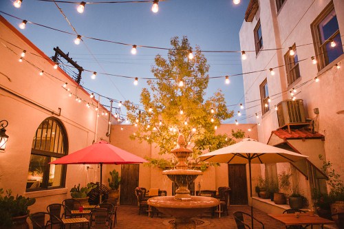 A cozy courtyard with a fountain, string lights, and shaded tables under umbrellas, creating a warm evening atmosphere.