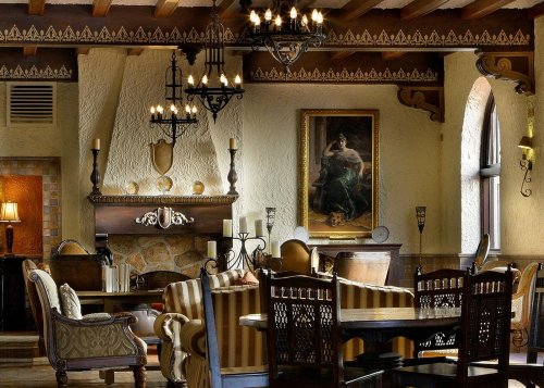 Elegant lounge with wooden beams, chandeliers, a fireplace, and plush seating, featuring a large portrait on the wall.