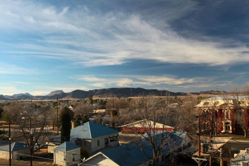 A scenic view of a small town with houses, trees, and mountains under a blue sky with wispy clouds.