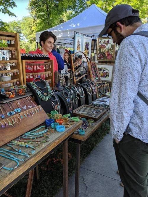 A vendor smiles at a customer browsing colorful jewelry and accessories at an outdoor market.