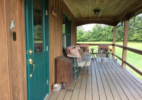 A cozy porch with a green door, seating, and a table, surrounded by greenery and a peaceful outdoor view.