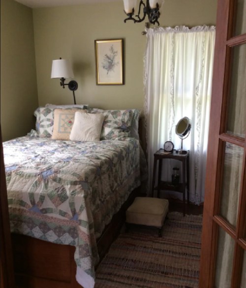 Cozy bedroom with a quilted bed, a lamp, a mirror on a nightstand, and a window with sheer curtains.
