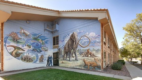 Mural on a building featuring prehistoric animals like dinosaurs, an elephant, and a turtle, with two figures in front.