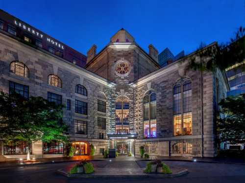 Historic building at night, featuring large windows and a decorative clock, surrounded by greenery and city lights.