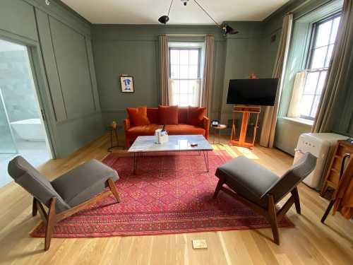 Cozy living room with an orange sofa, gray chairs, a coffee table, and a patterned rug, featuring green walls and natural light.