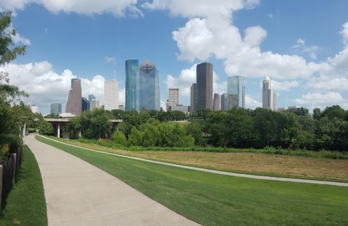 Panoramic view of a city skyline with tall buildings, green parkland, and a blue sky with fluffy clouds.