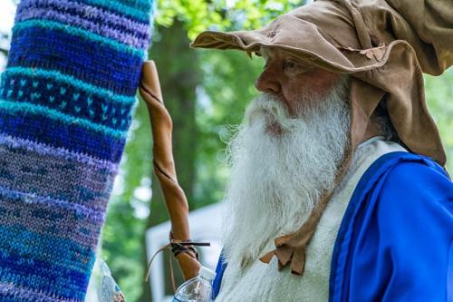 A bearded man in a wizard costume with a large hat and blue robe, standing next to a colorful, knitted pole.