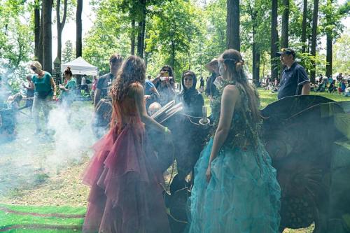 A group of people in colorful costumes gather in a forested area, with smoke and tents in the background.