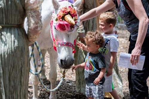 Two children pet a unicorn adorned with flowers and a horn, while adults stand nearby in a sunny outdoor setting.