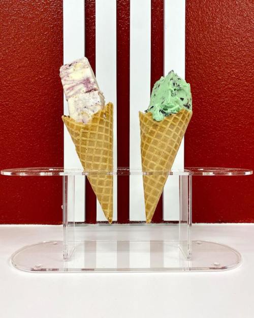 Two ice cream cones in a clear holder, one with purple swirls and the other mint green, against a red wall.