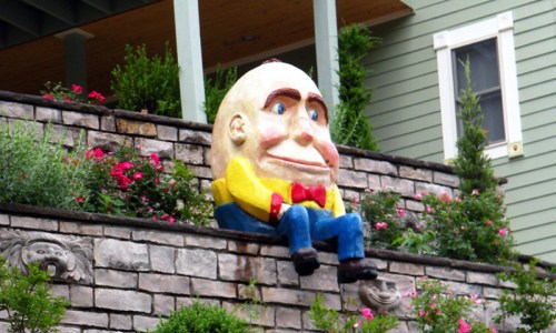 A large, cartoonish statue of a smiling man with a bow tie sits on a stone wall surrounded by colorful flowers.