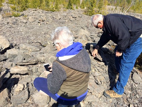 Two people explore a rocky area, one sitting and using a phone, while the other examines the ground.