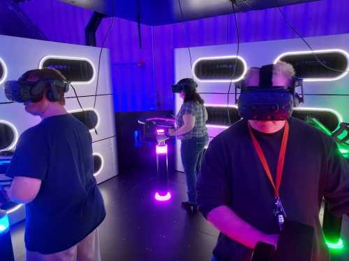 Three people wearing VR headsets in a colorful gaming room, interacting with virtual elements.