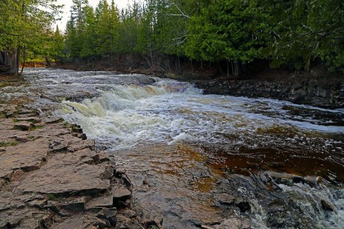 A serene river flows over rocky terrain, surrounded by lush green trees and a peaceful natural landscape.